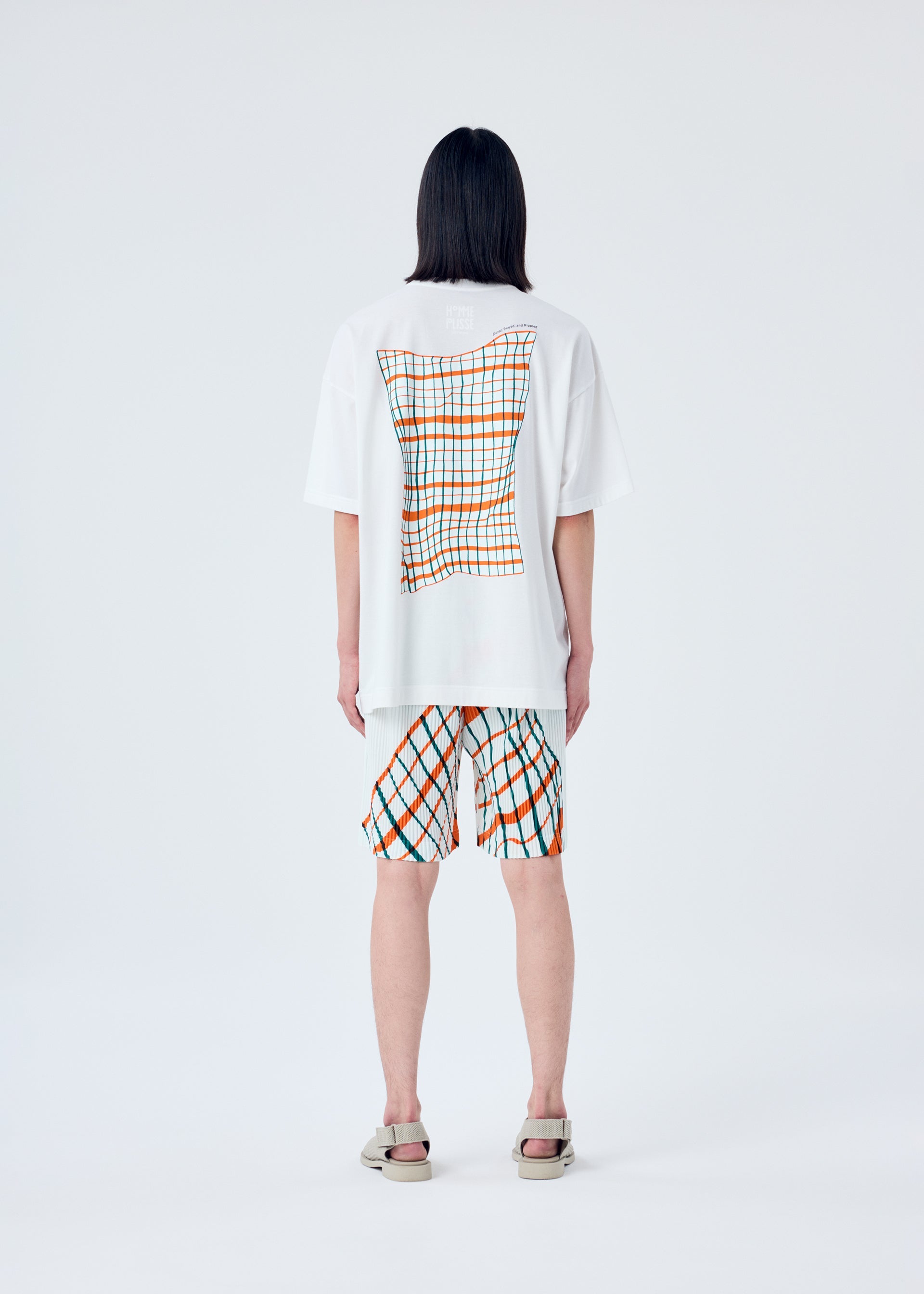 A model wears the HOMME PLISSÉ ISSEY MIYAKE WINDSWEPT PLAID trousers.