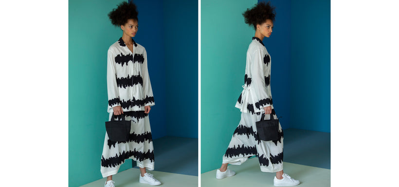 Left: Model wearing WAVY BORDER Shirt and Trousers in WhitexBlack, standing against a backdrop of green and blue wall panels. Right: Model wearing WAVY BORDER Shirt and Trousers in WhitexBlack, captured mid-stride in a dynamic wide-step pose, set against panels of green and blue.