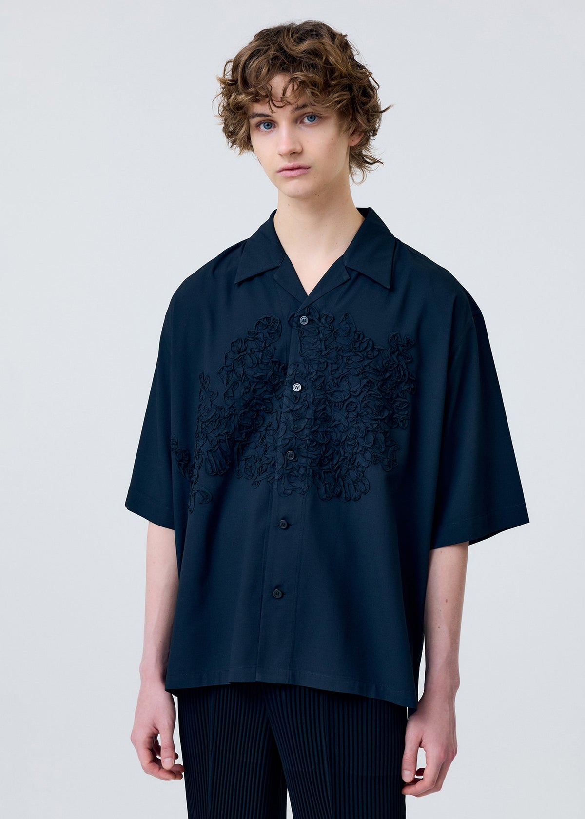A model wears the HOMME PLISSÉ ISSEY MIYAKE EMBROIDERY PRESS SHIRT shirt.