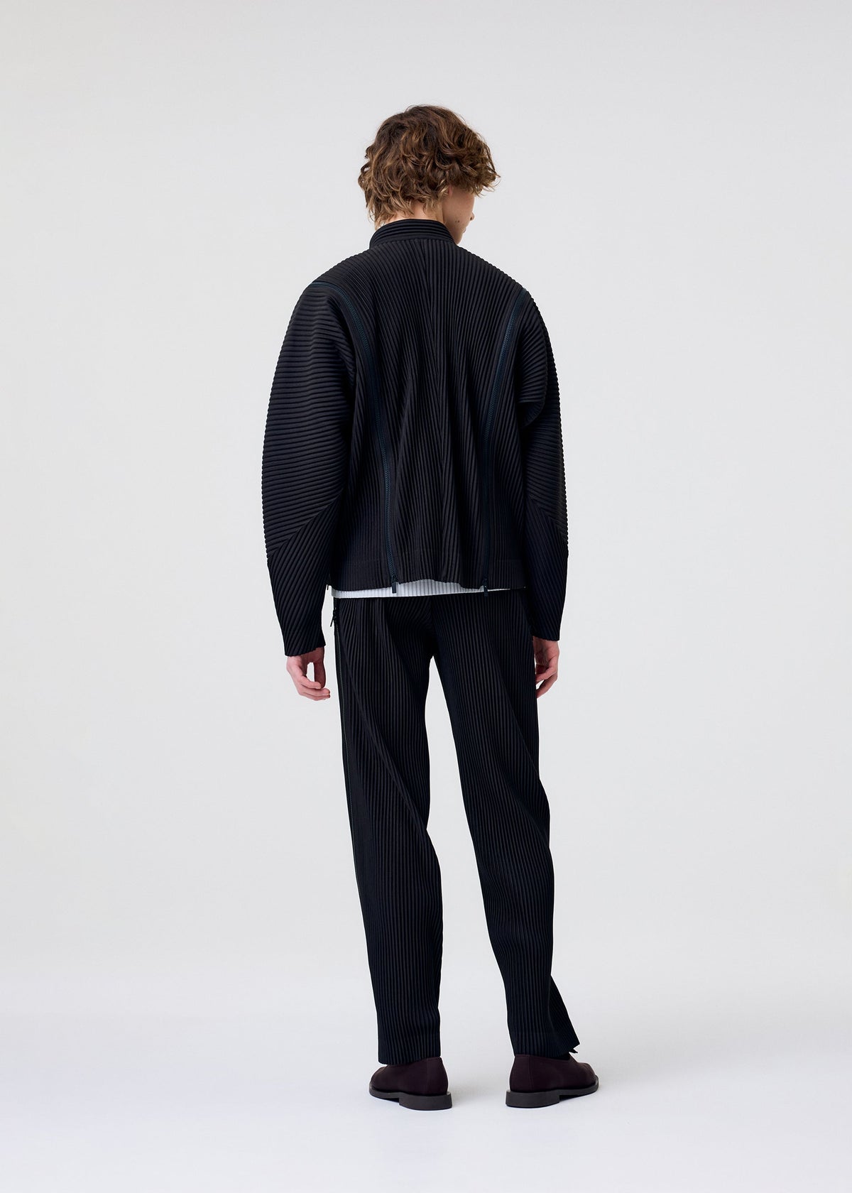 A model wears the HOMME PLISSÉ ISSEY MIYAKE MULTI-FASTENED multi-fastened-trousers-pizza-scorch-brown.