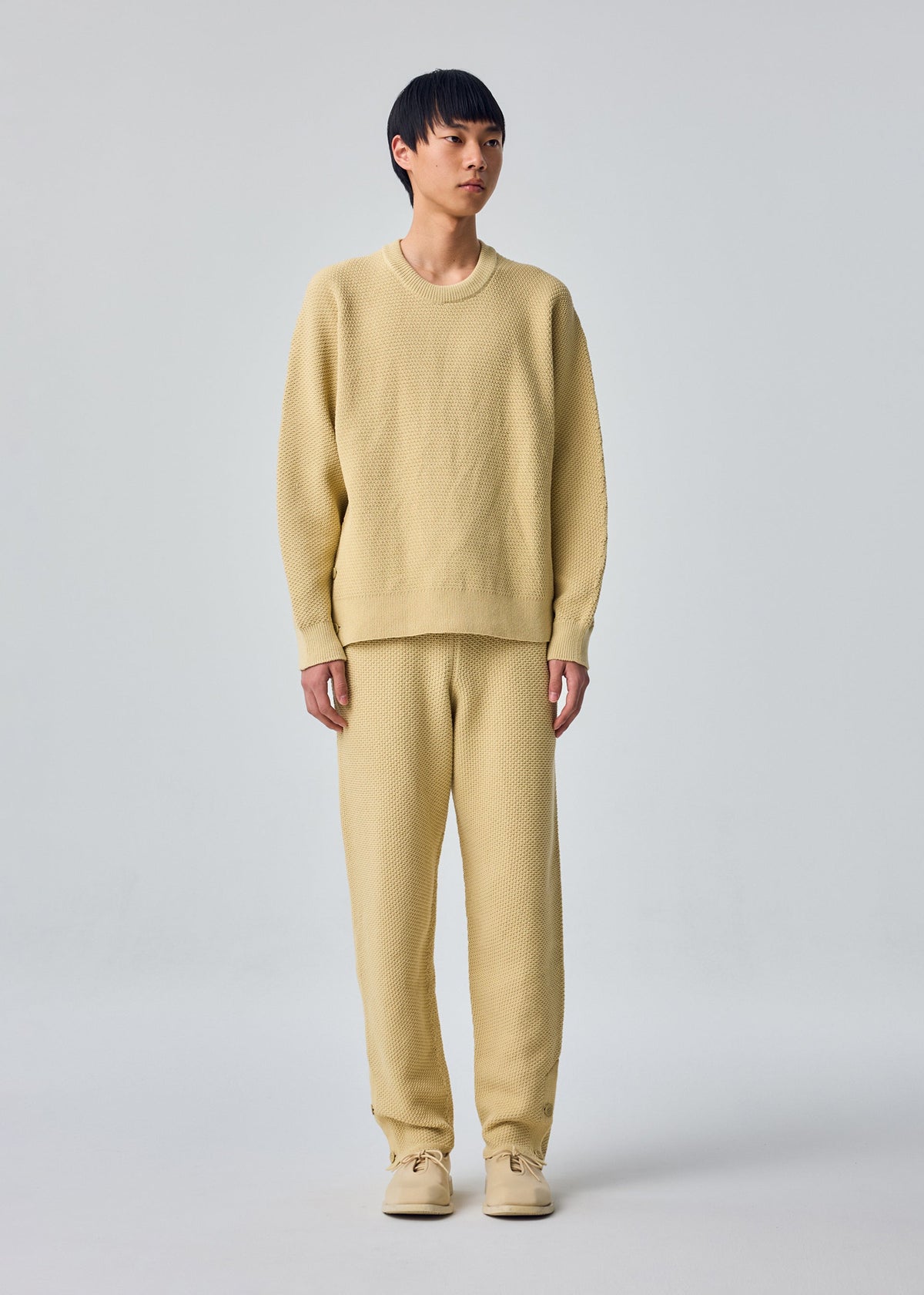 A model wears the HOMME PLISSÉ ISSEY MIYAKE RUSTIC KNIT rustic-knit-top-yellow-beige.