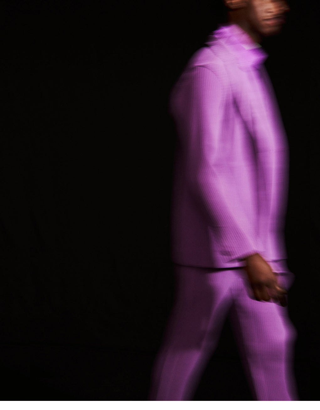 Model mid walking, dressed in a bright purple pleated suit over a purple shirt, shot in front of a black background. They're visible from their ankles to their eyebrows with the right side facing the camera.