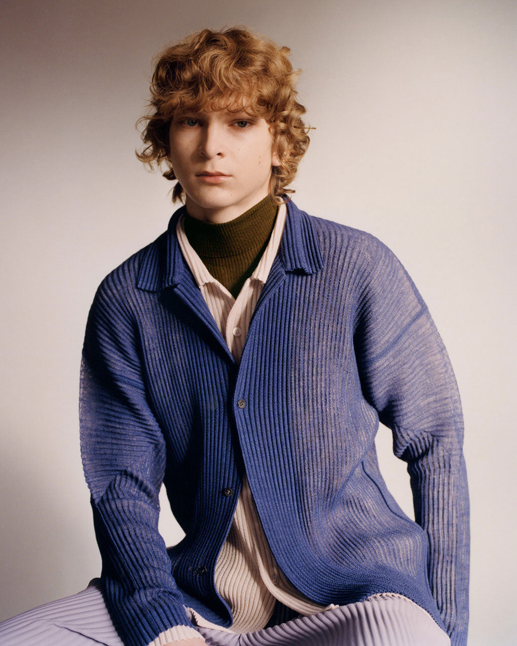 Model sat down, wearing a pleated blue shirt layered over a beige shirt over a brown turtleneck top paired with light purple pleated trousers. Posed in front of a beige background.