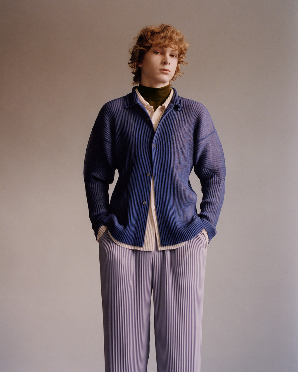 Standing model wearing a pleated blue shirt layered over a beige shirt over a brown turtleneck top paired with light purple pleated trousers. Posing with hands in their trouser pockets in front of a beige background.