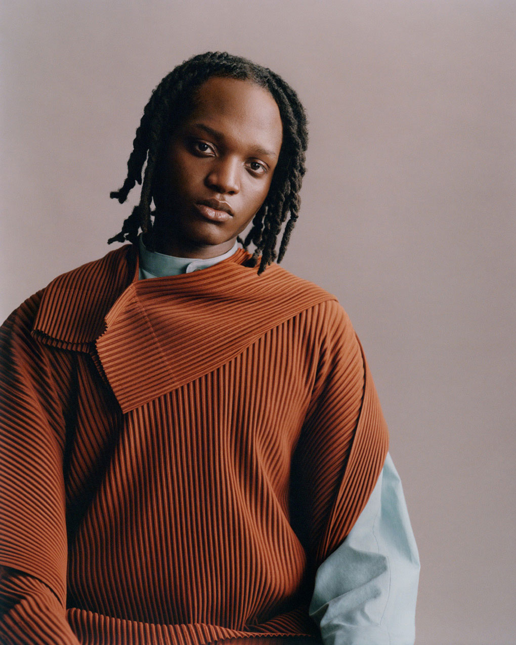 Model wearing terracotta pleated top with one long sleeve and one short over a pale blue shirt. Posing in front of a pale beige background. Shot from hips up.