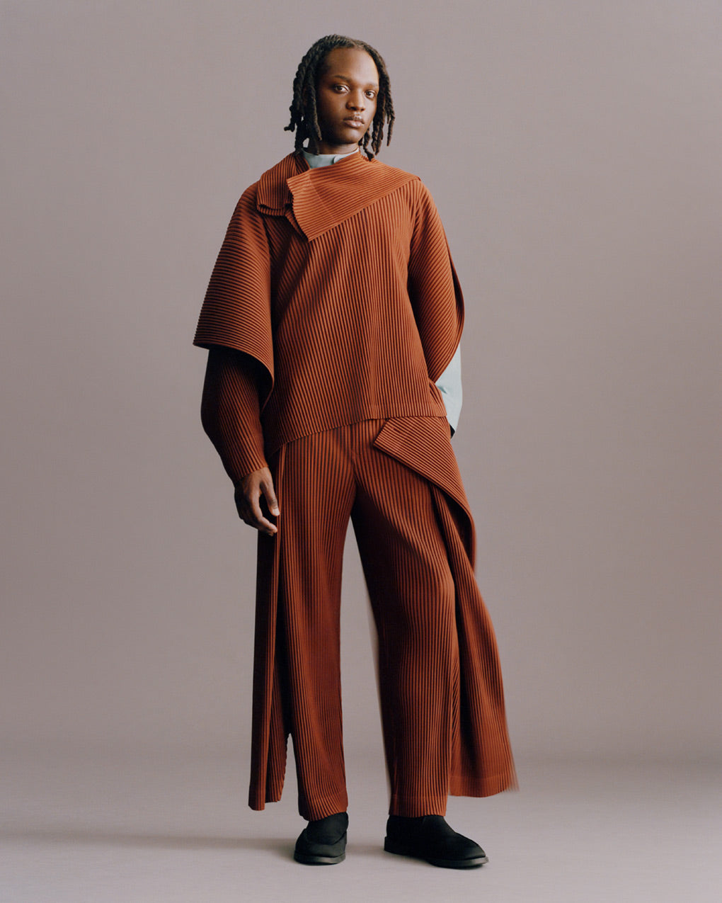 Model wearing terracotta pleated top with one long sleeve and one short over a pale blue shirt, paired with matching terracotta wide-leg pleated trousers and black shoes. Posing in front of a pale beige background. 