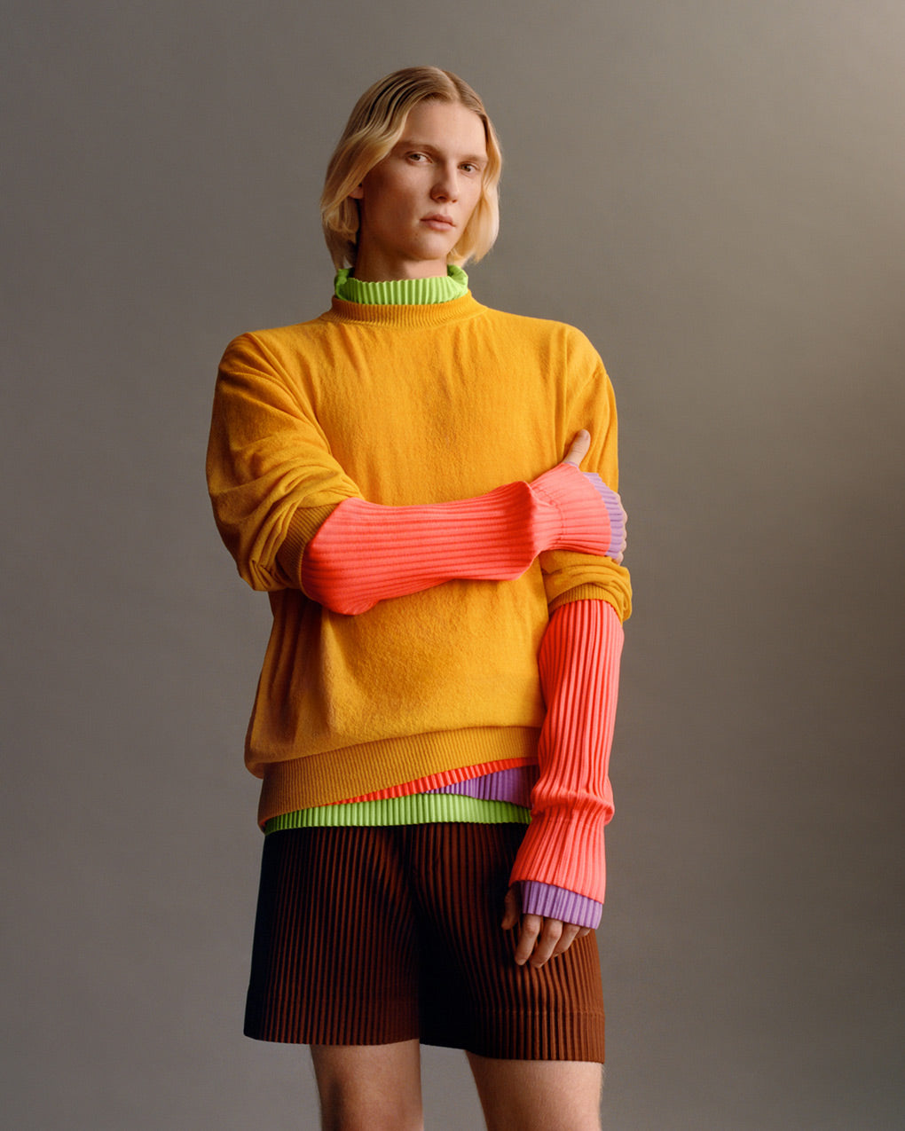 Standing model wearing brown pleated shorts and a mustard knit jumper over layers of pleated tops; one light green, one purple and one coral pink. Posing in front of a light grey background, shot from knees up.