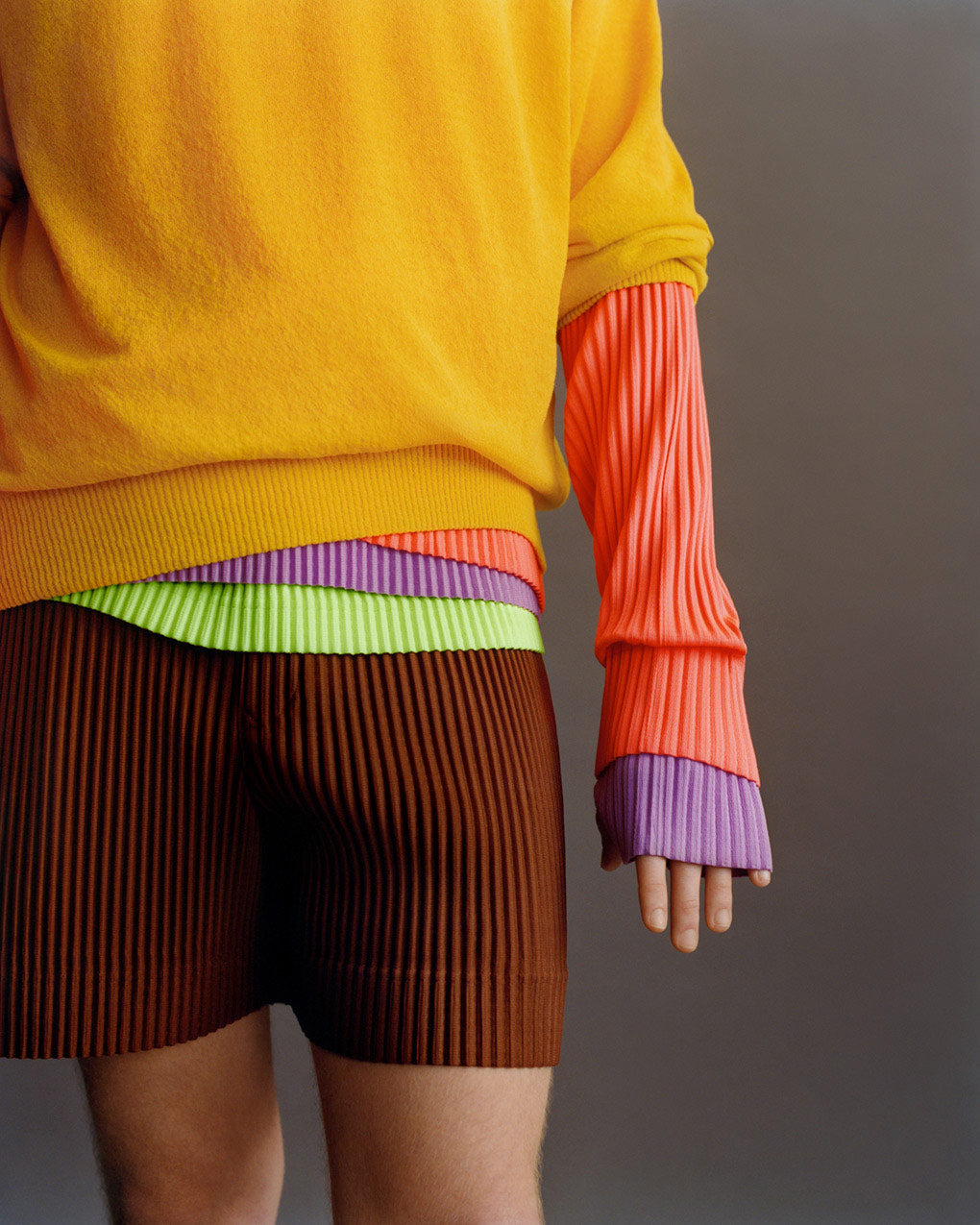 Closeup of model wearing brown pleated shorts and a mustard knit jumper over layers of pleated tops; one light green, one purple and one coral pink. Posing in front of a light grey background, visible from knees to chest.