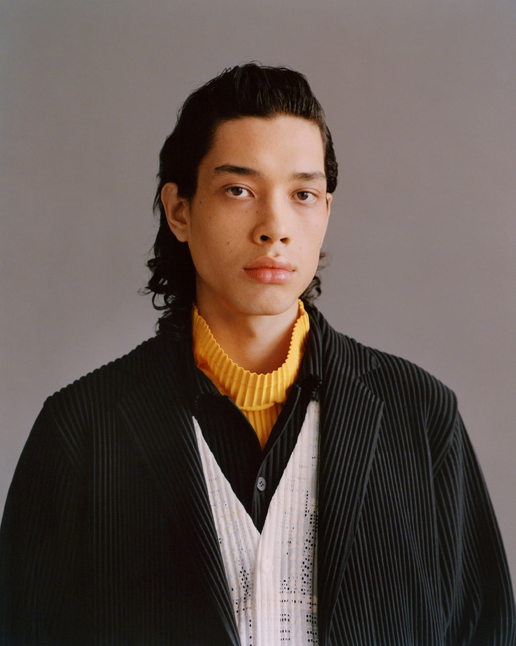 Portrait of model wearing dark green pleated jacket over a white pleated cardigan, black pleated shirt and a yellow pleated turtleneck top. Posed in front of a light grey background.