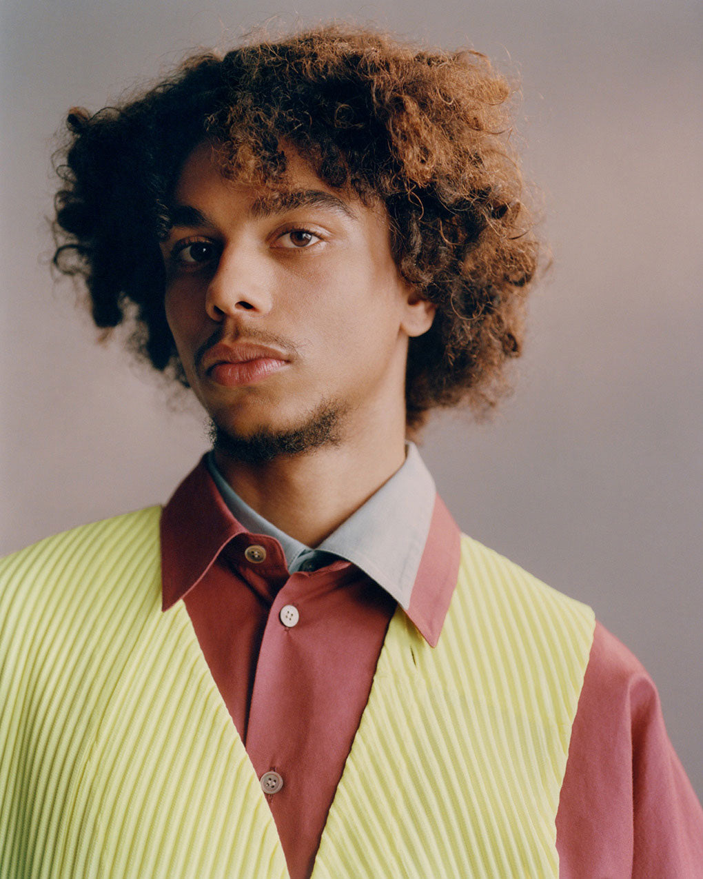 Portrait of model wearing a bright yellow green pleated vest over a red shirt layered ontop of a light grey shirt. Posed in front of a light grey background.