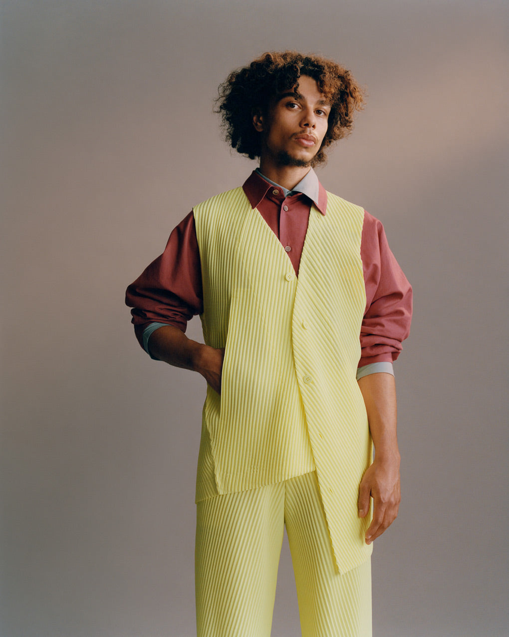 Portrait of model wearing a bright yellow green pleated vest over a red shirt layered ontop of a light grey shirt. Paured with matching bright yellow green pleated trousers. Posed with right hand in vest pocket in front of a light grey background. Shot from knees up.
