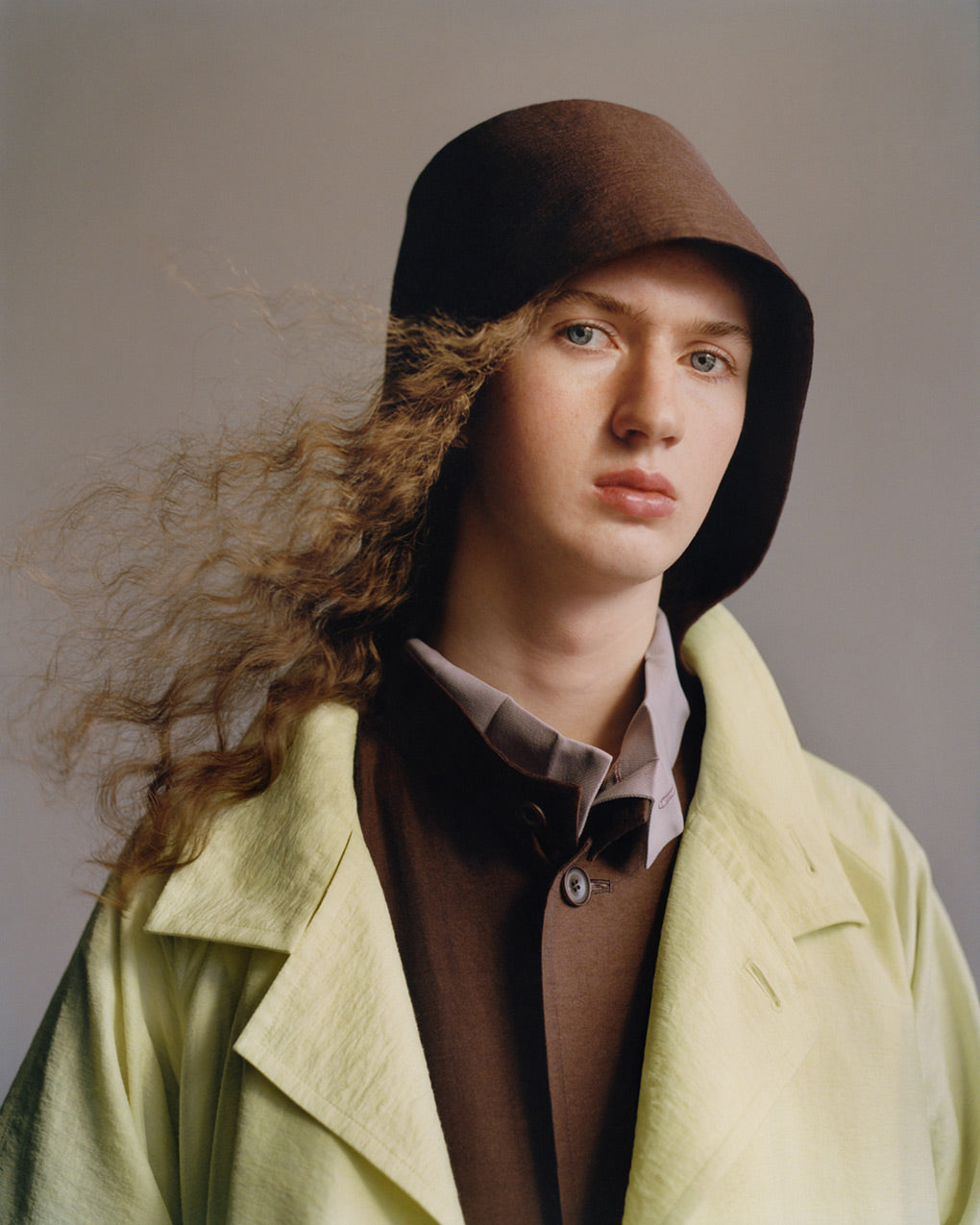 Portrait of model wearing a light green coat over a brown jacket with a hood which is worn up, layered ontop of a light grey shirt. Posed in front of a light grey background.