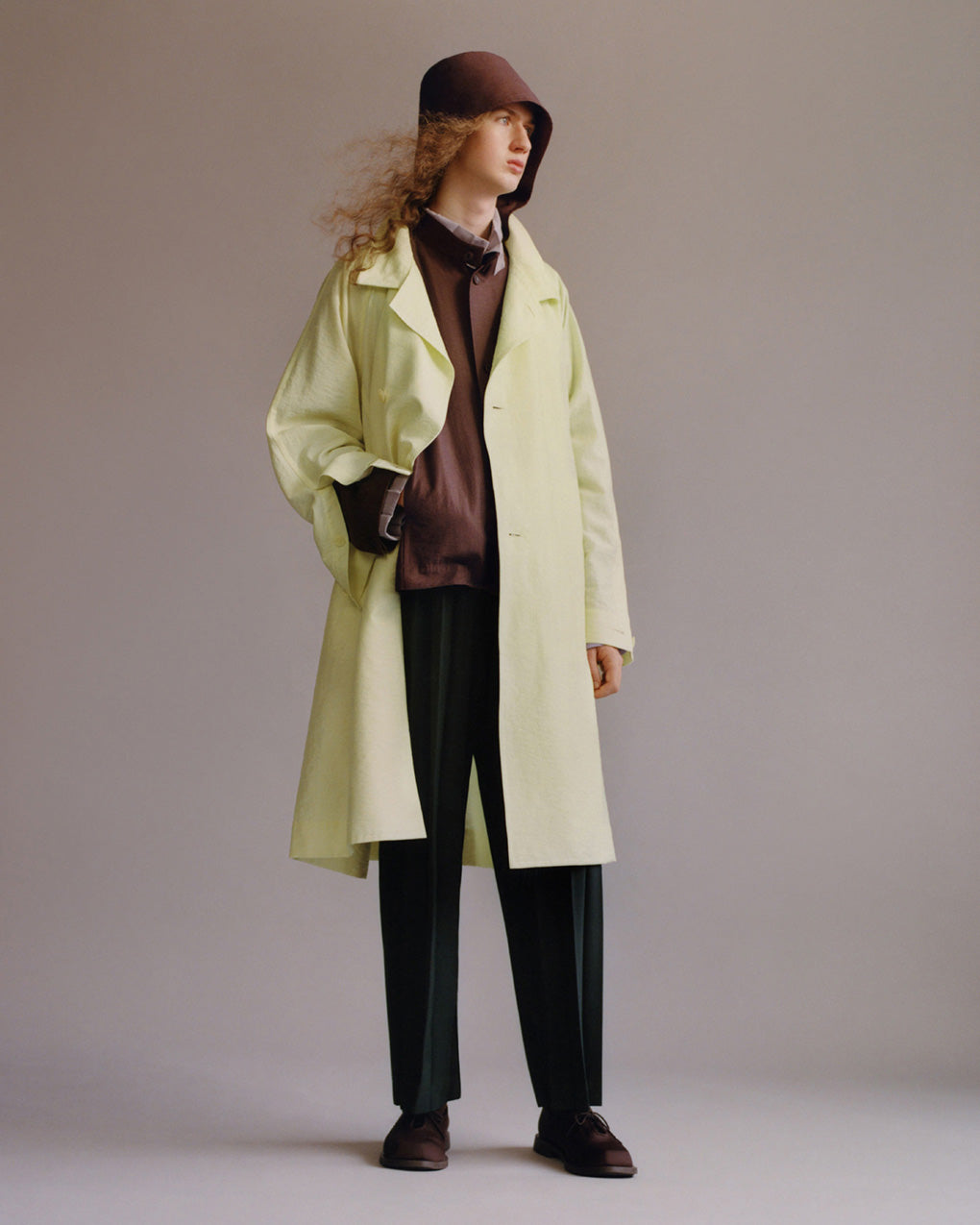 Standing model wearing a light green coat over a brown jacket with a hood which is worn up, layered ontop of a light grey shirt. Paired with black trousers and brown shoes. Posing with right hand in jacket pocket in front of a light grey background.