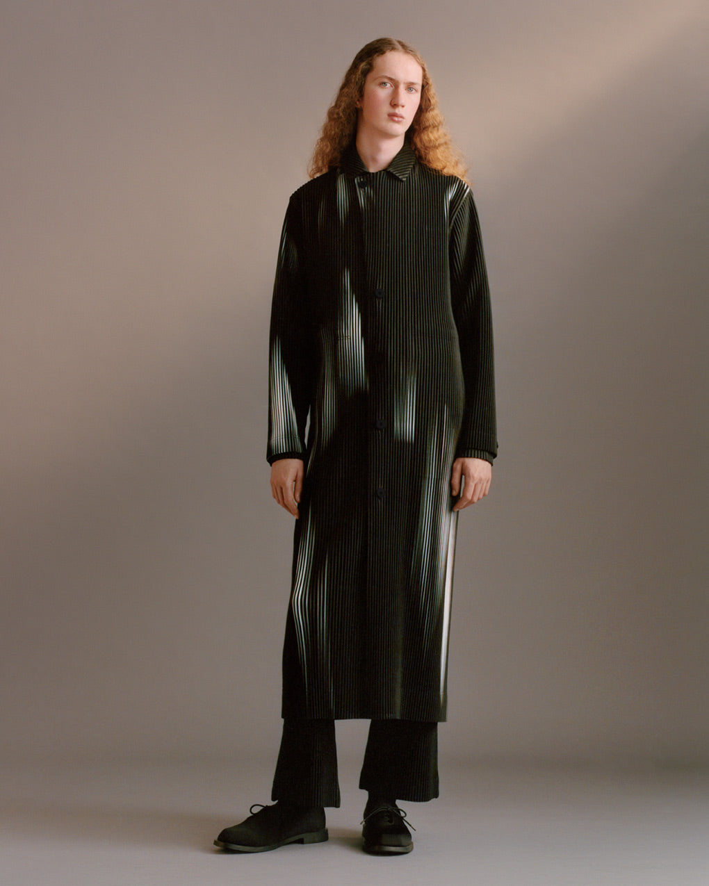 Standing model wearing black and white printed pleated long coat over black pleated trousers with black shoes. Piosing in front of a light grey background.