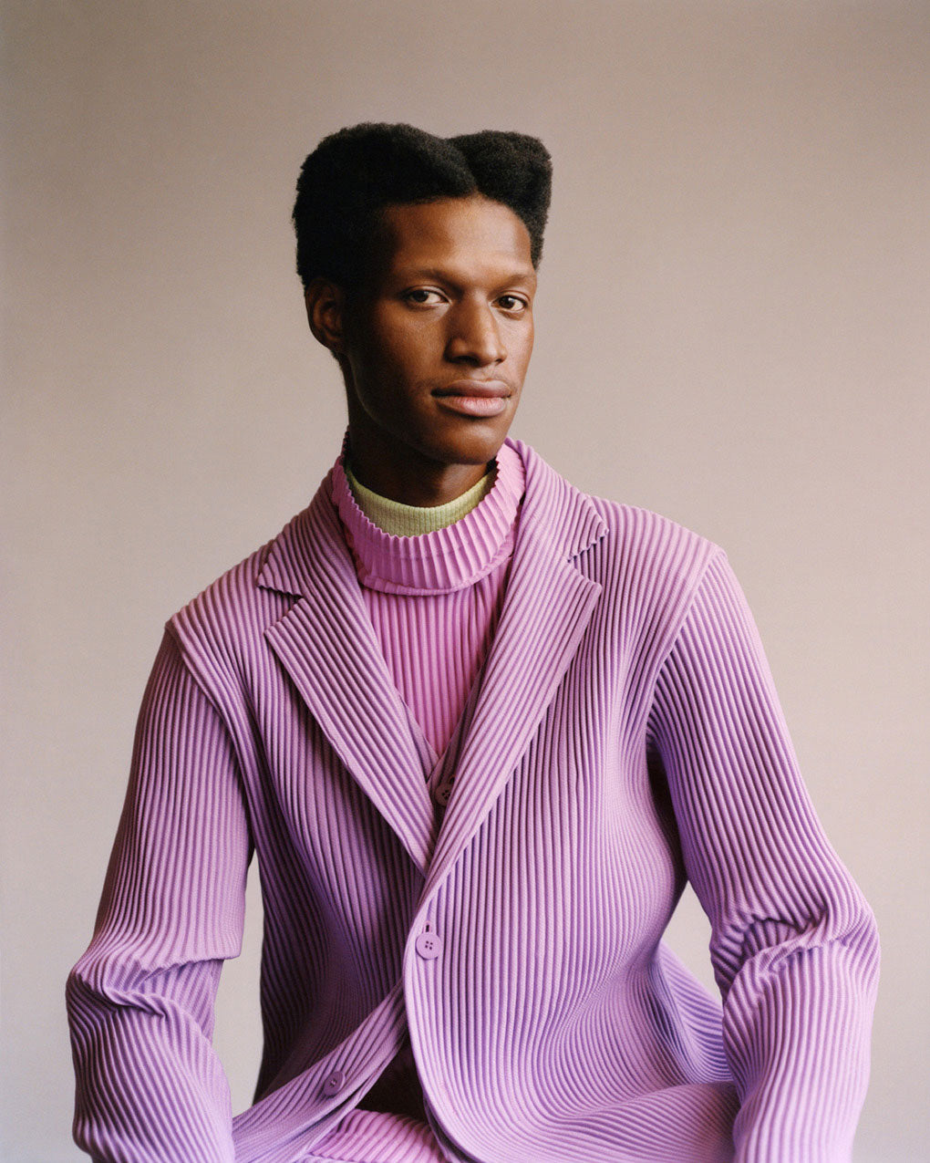 Portrait of model wearing a light purple suit with a light purple vest over a pink turtleneck top over a yellow turtleneck top. Posing in front of a light grey background.