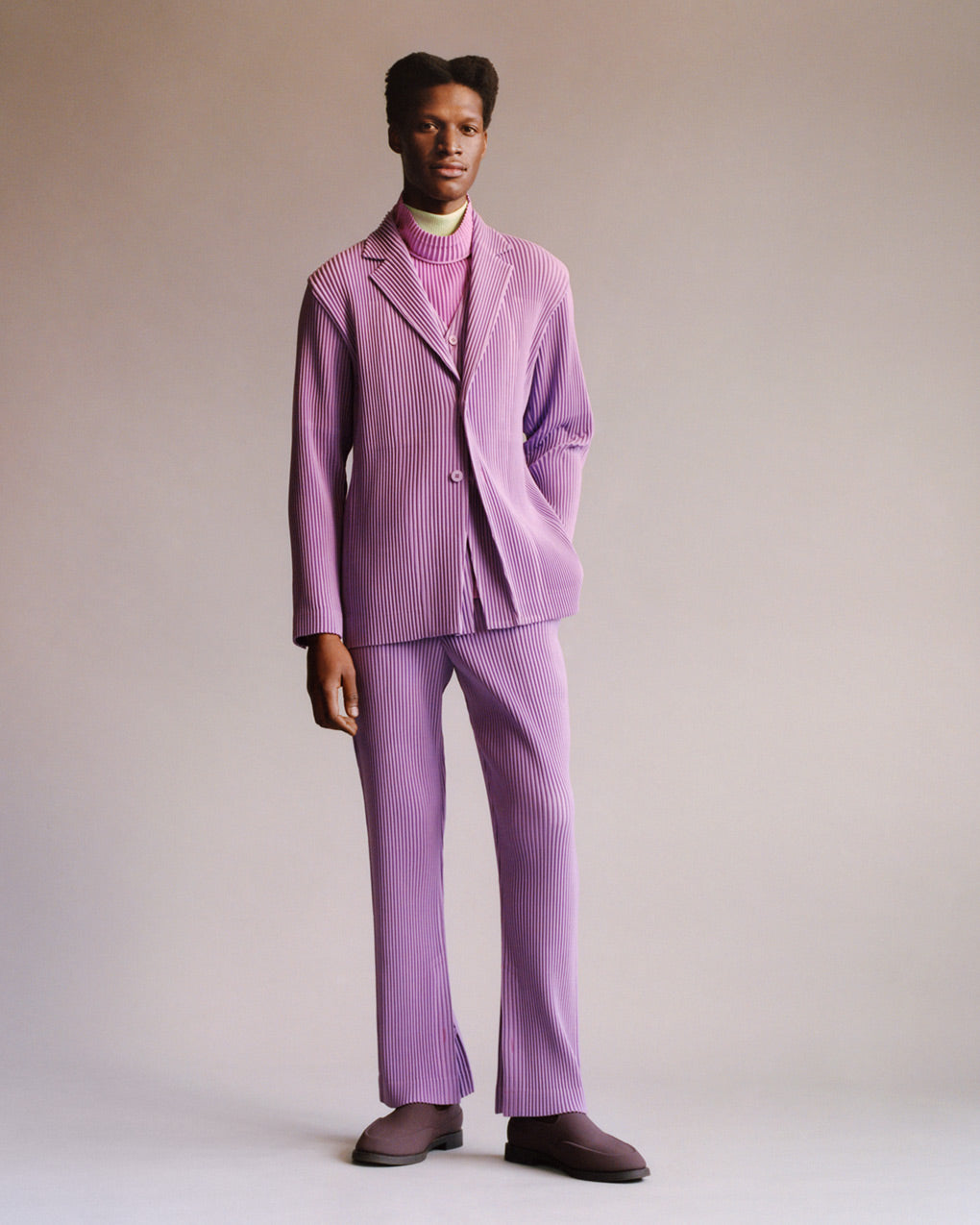 Standing model wearing a light purple suit with a light purple vest over a pink turtleneck top over a yellow turtleneck top and brown shoes. Posing with left hand in trouser pocket in front of a light grey background.