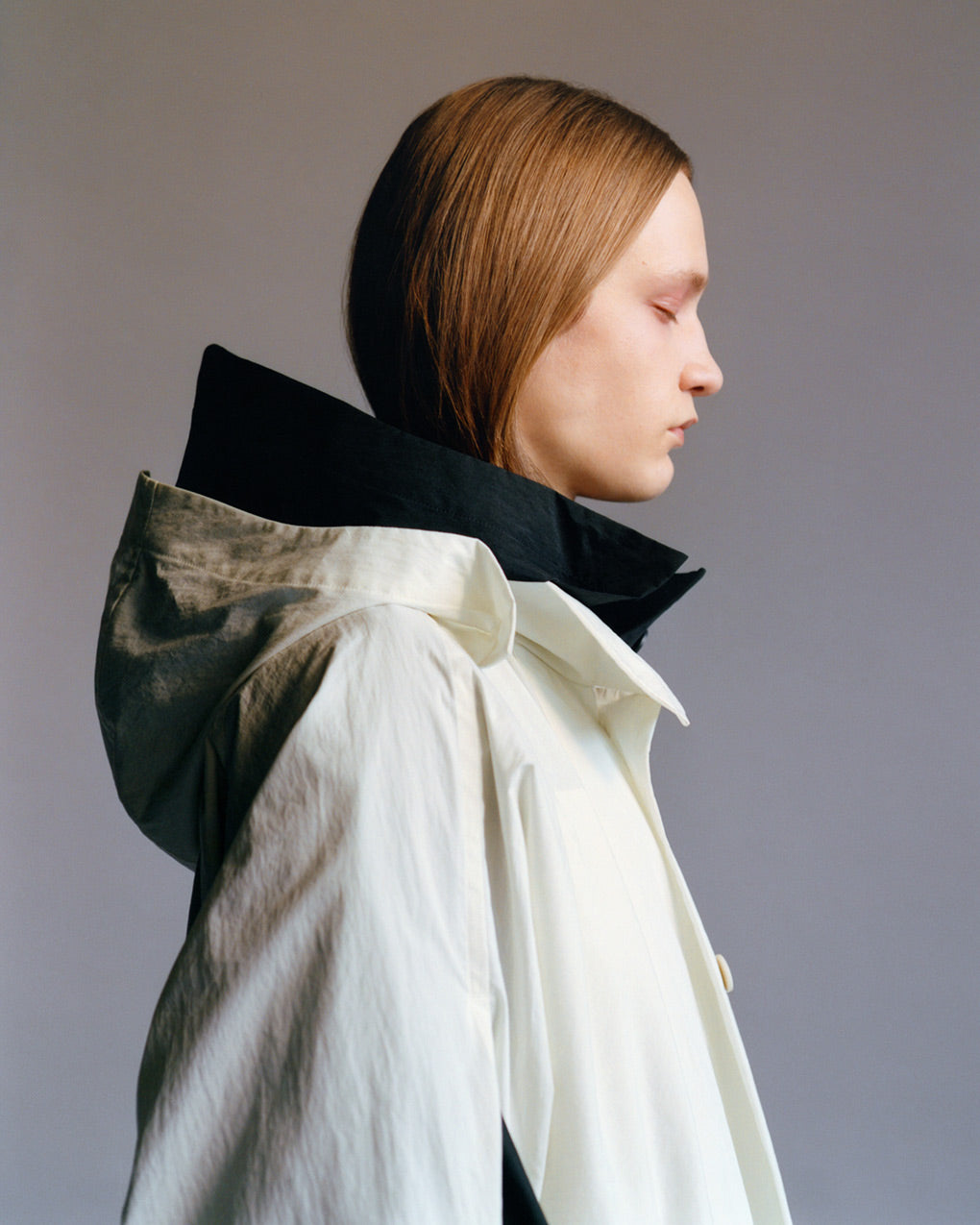 Profile portrait of a model wearing a white coat with a hood over a black coat with a hood, both hoods are down. Posed in front of a light grey background.