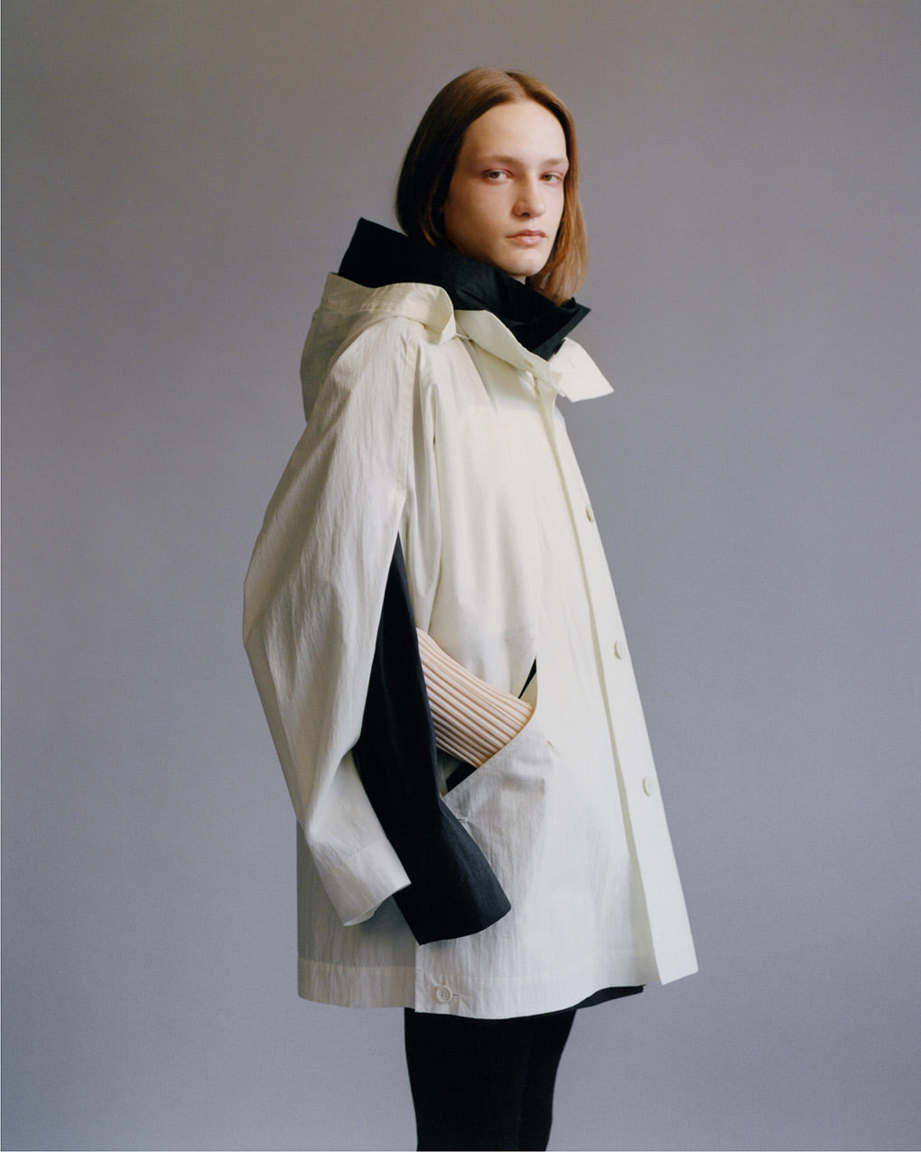 Standing model wearing a white coat with a hood over a black coat with a hood, both hoods are down. Worn over a beige long-sleeved pleated top and black pleated trousers. Posed in front of a light grey background and shot from knees up.