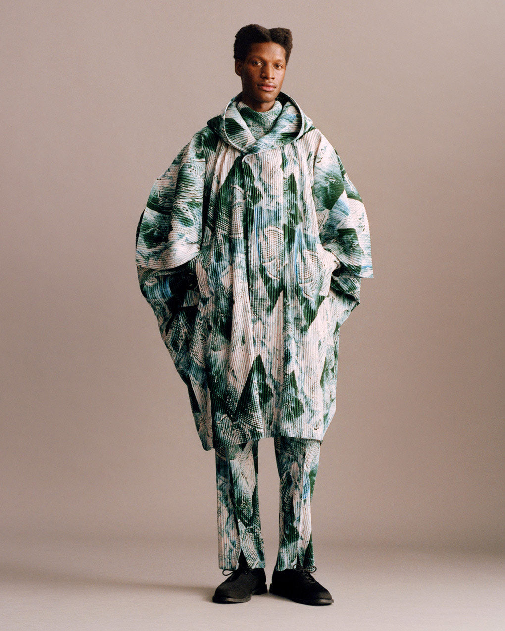 Standing model wearing pleated coat and trousers in a busy print with shades of white, blue and green. Paired with black shoes. Posing with hands in coat pocets in front of a light beige background.