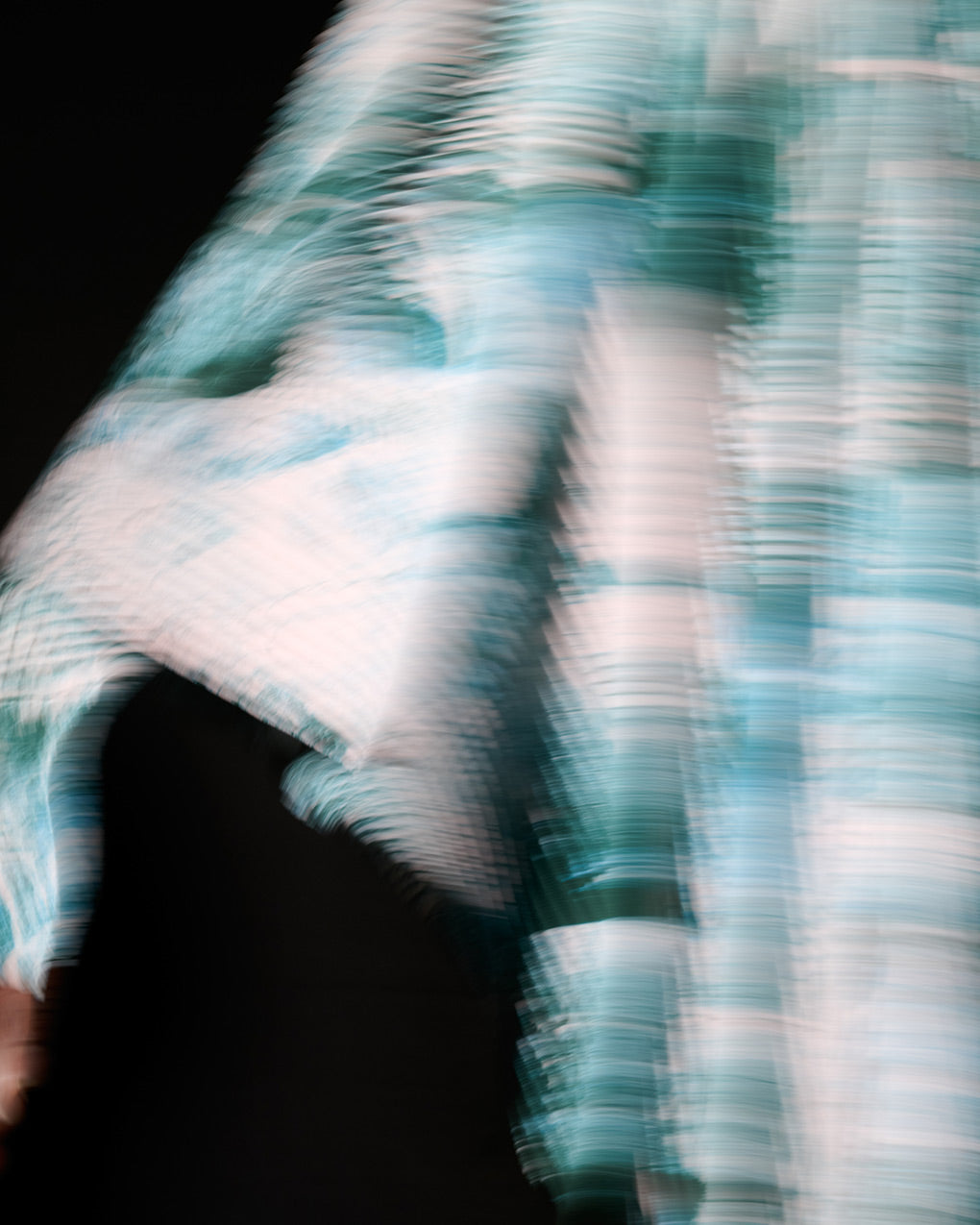 Closeup of model wearing pleated coat in a busy print with shades of white, blue and green. Moving in front of a black background.