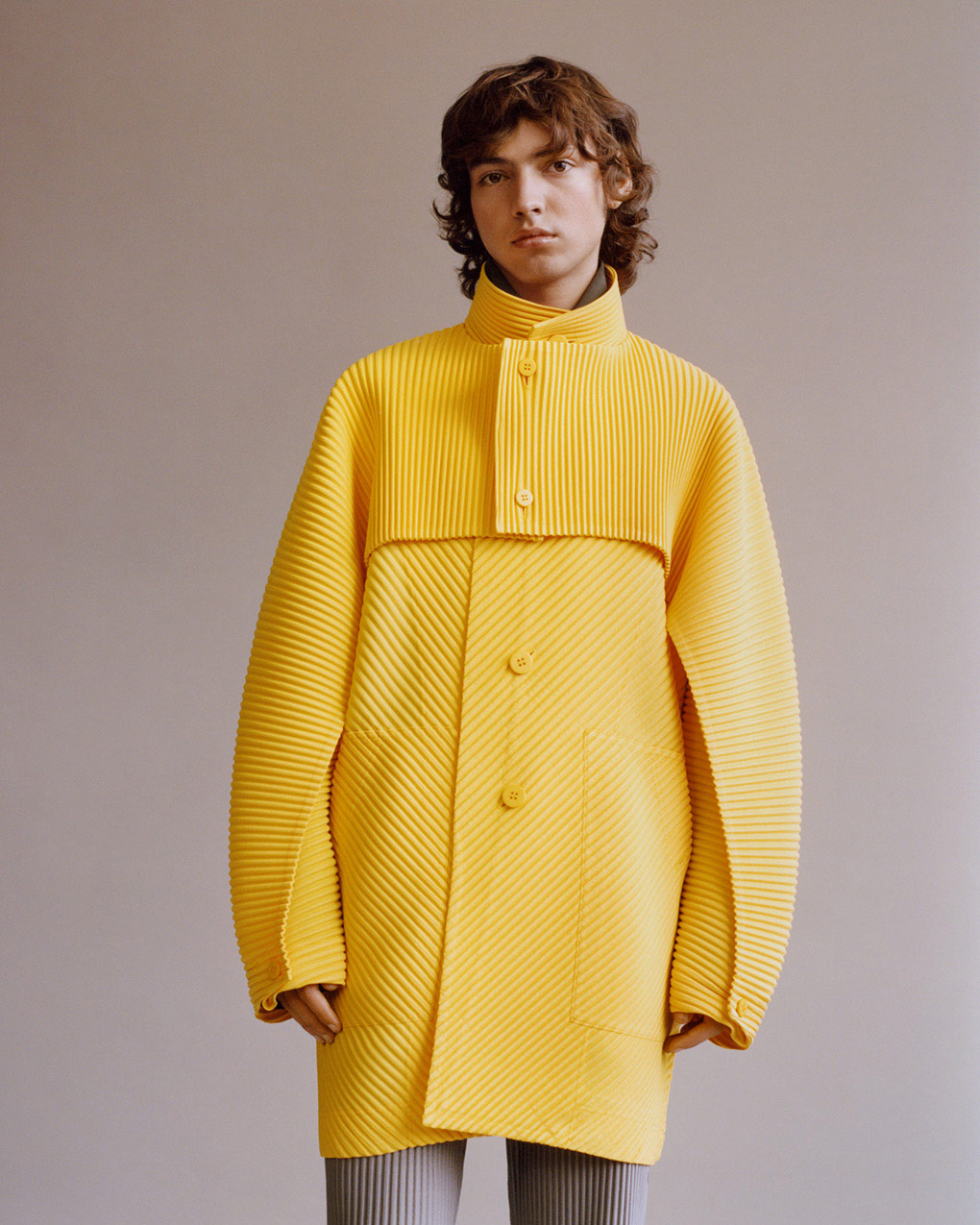 Standing model wearing a bright yellow long-sleeved pleated boler over a bright yellow pleated coat with a high neckline over a brown shirt. Paired with light grey  pleated trousers. Posing in front of a light grey background. Shot from knees up. 