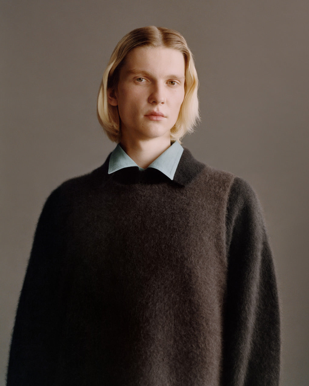 Portrait of model wearing wooly brown vest over a black wooly shirt over a pale blue shirt. Posing in front of a dark grey background.