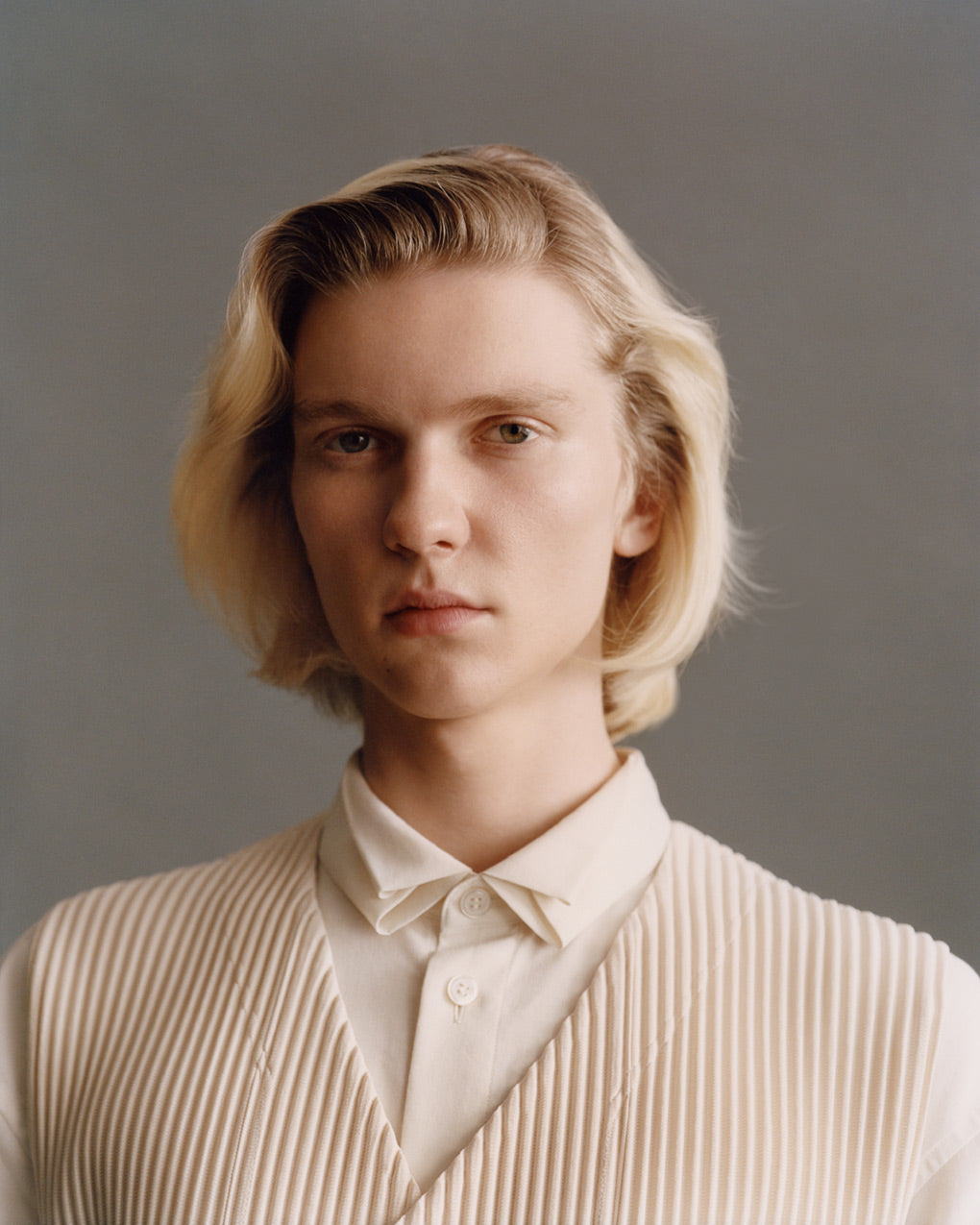 Portrait of model wearing beige pleated vest over a white shirt. Posed in front of a dark grey background.
