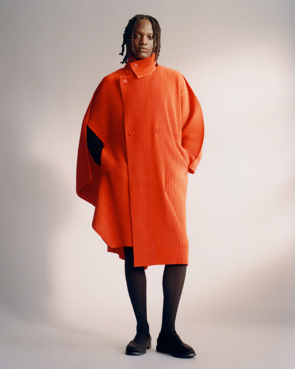 Standing model wearing a bright red pleated coat over black tights with black shoes. Posed in front of a light grey background with hands in coat pockets.