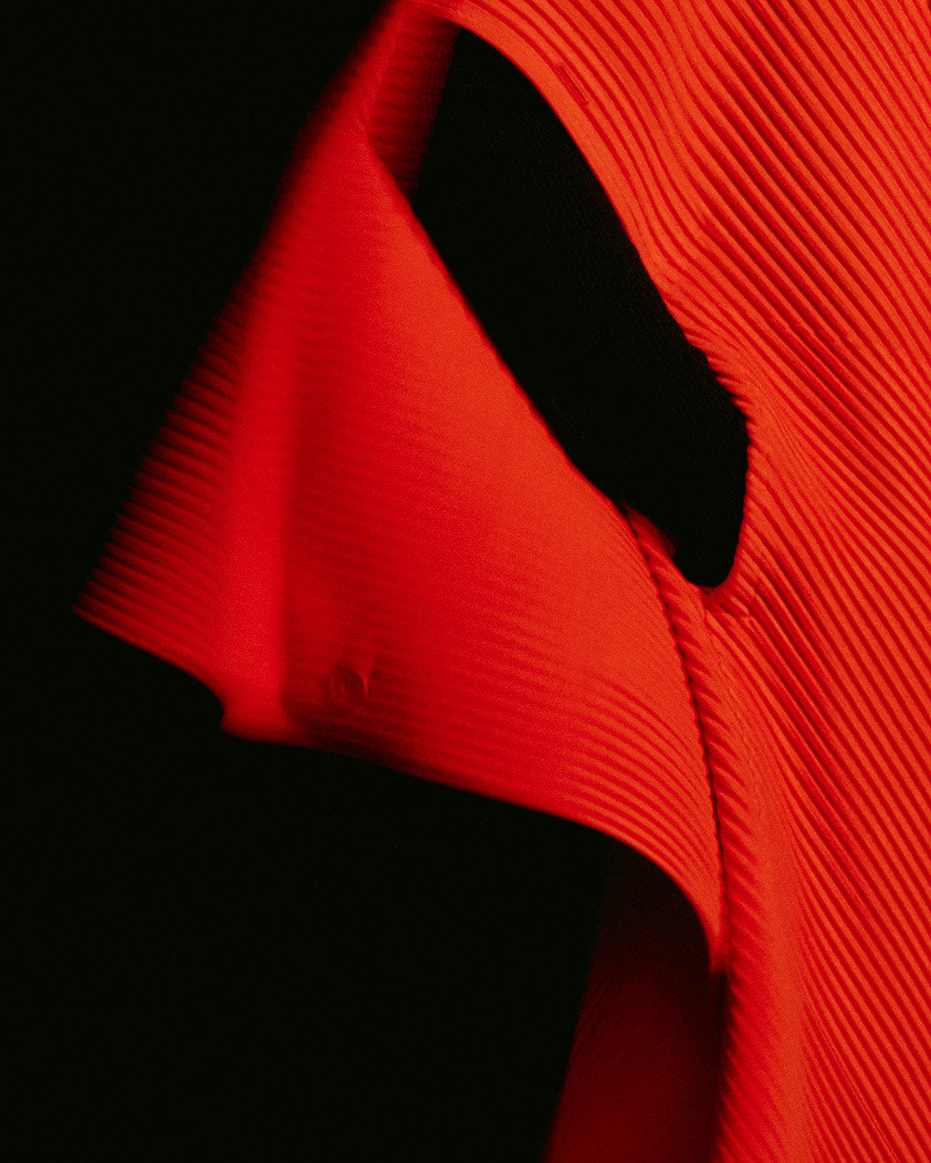 Closeup of model wearing a bright red pleated coat. Black background.