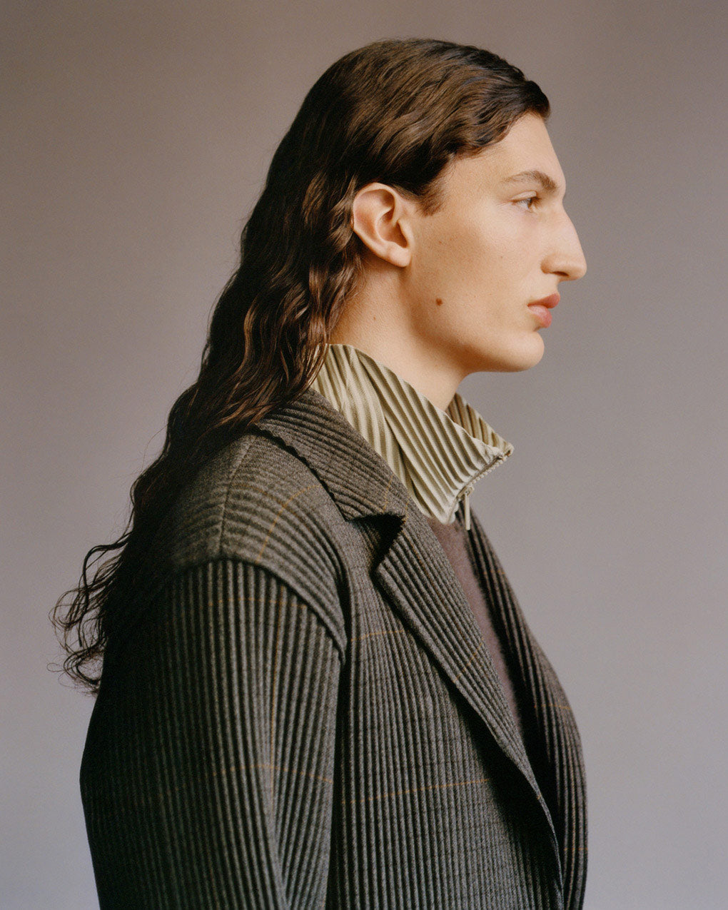 Profile portrait of model wearin ga pleated jacket with a tweed pattern over a wooly brown vest and a grey beige pleated zip up jacket with a stand collar. Posing in front of a grey background.
