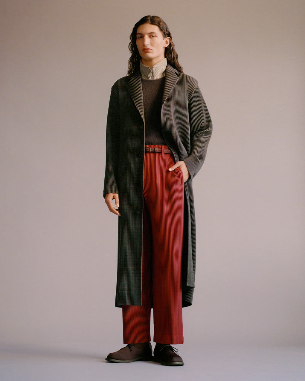Standing model wearing a pleated long jacket with a tweed pattern over a wooly brown vest and a grey beige pleated zip up jacket with a stand collar. Paired with red trousers, a brown belt and brown shoes. Posing with left hand in trouser pocket in front of a grey background.