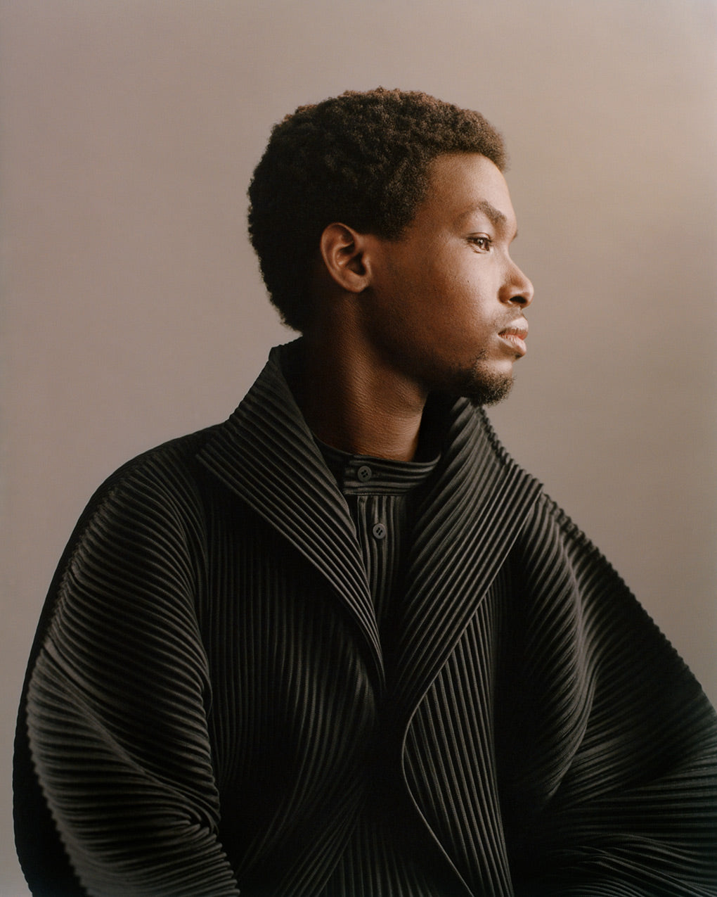 Profile portrait of model wearing a black pleated coat over a black pleated shirt. Posing in front of a grey background.