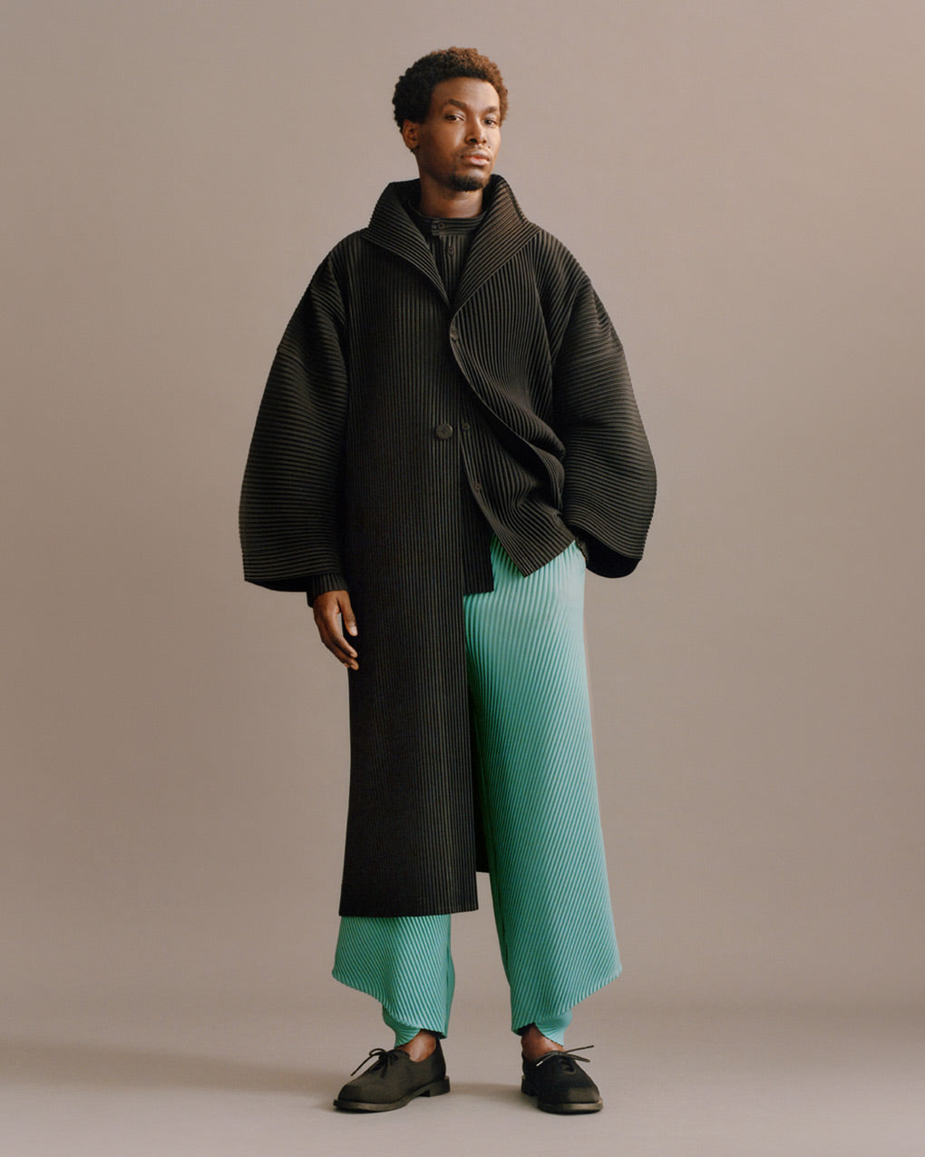 Standing model wearing a black pleated coat over a black pleated shirt. Paired with turquoise blue pleated trousers and black shoes. Posing with left hand in trouser pocket in front of a grey background.