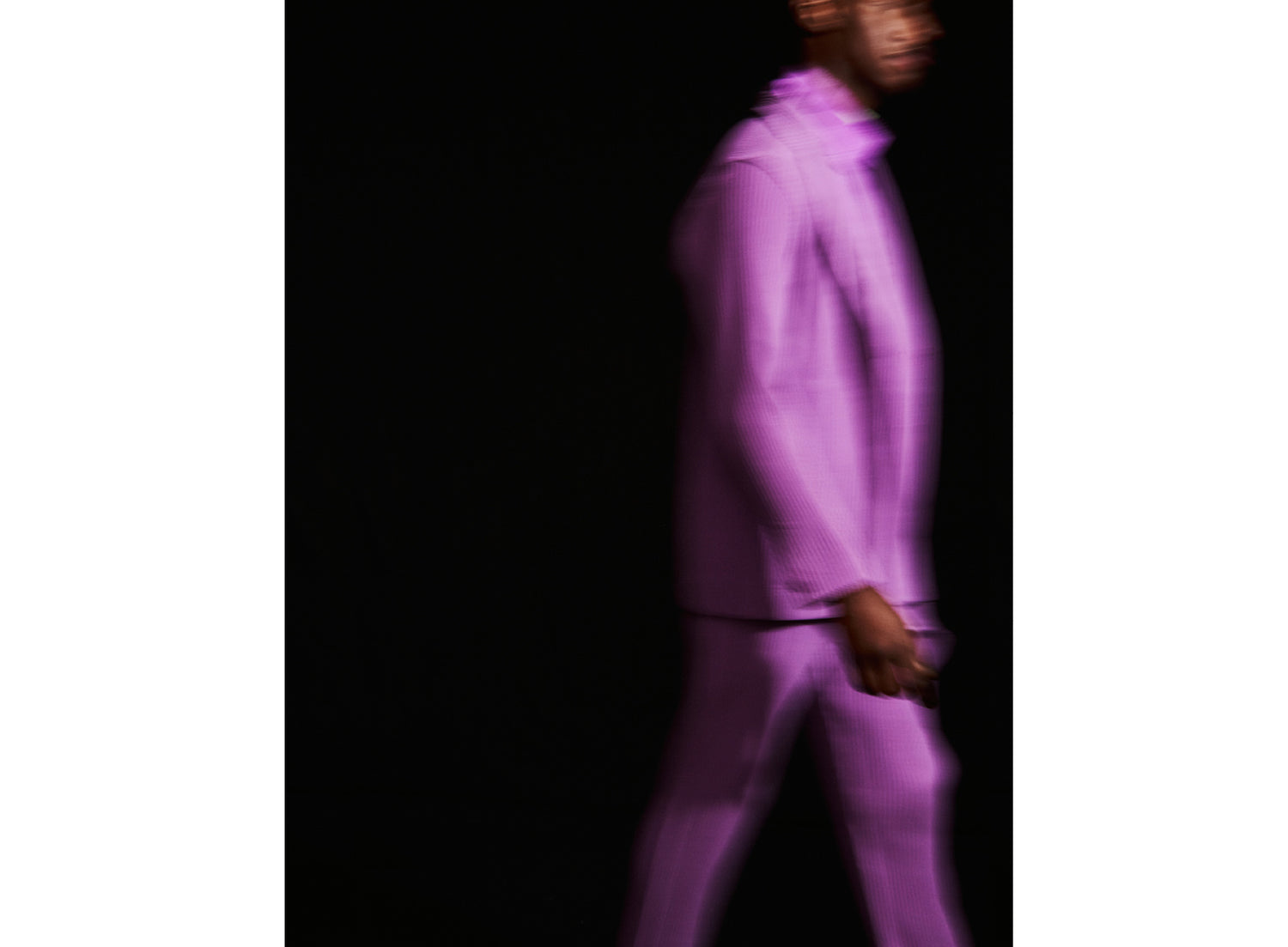 Model mid walking, dressed in a bright purple pleated suit over a purple shirt, shot in front of a black background. They're visible from their ankles to their eyebrows with the right side facing the camera.