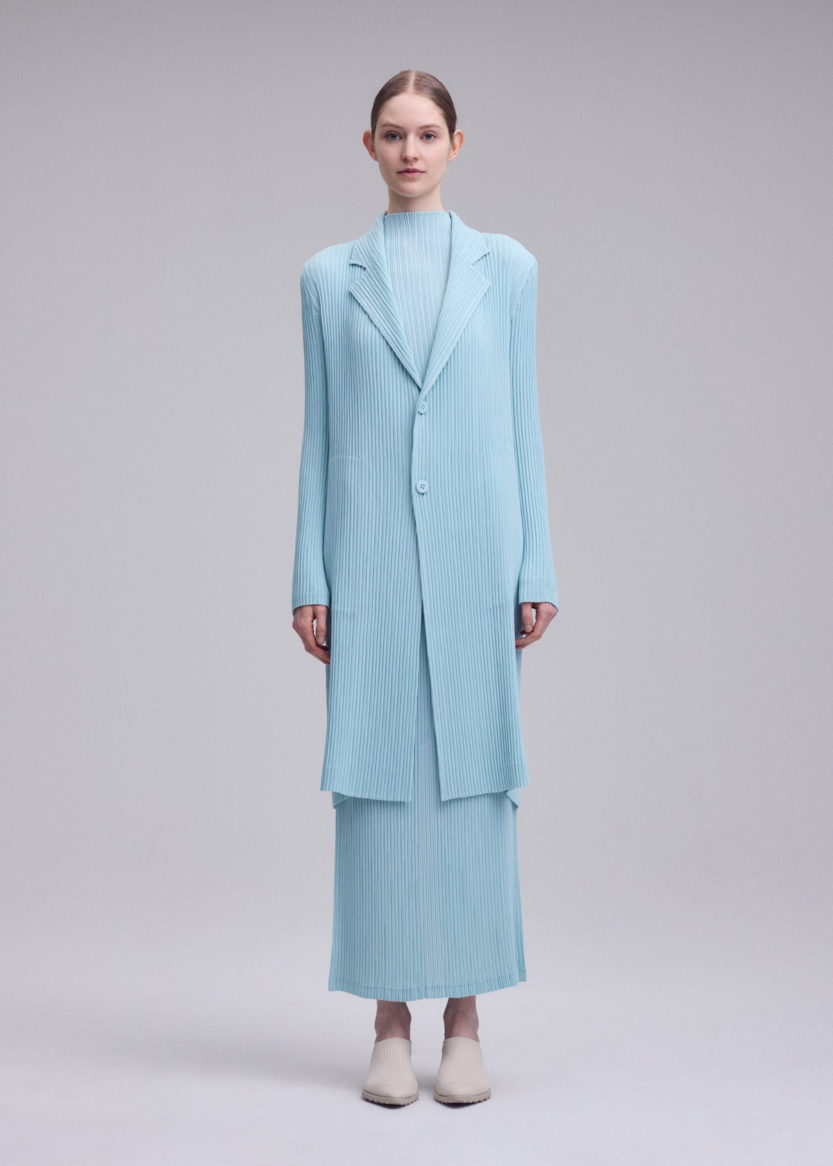 A model wears the ISSEY MIYAKE HATCHING PLEATS hatching-pleats-three-quarter-length-coat-grey.