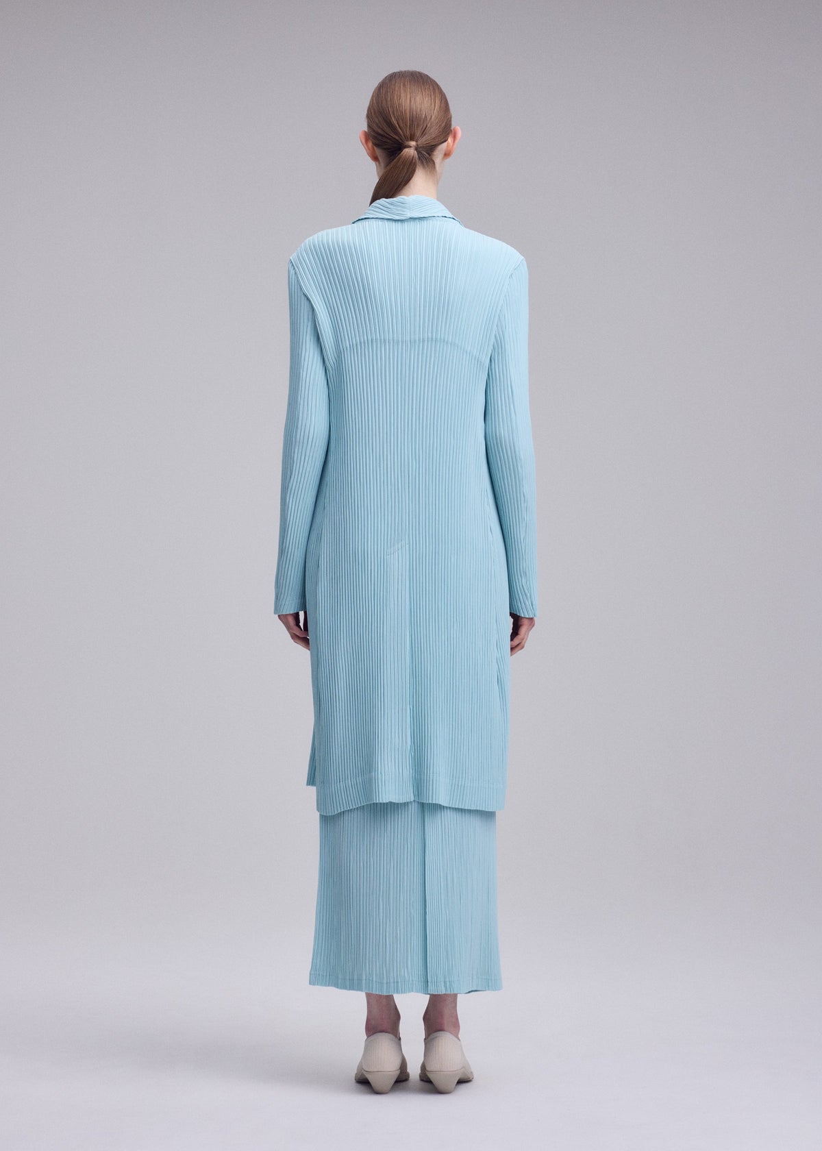 A model wears the ISSEY MIYAKE HATCHING PLEATS hatching-pleats-three-quarter-length-coat-grey.