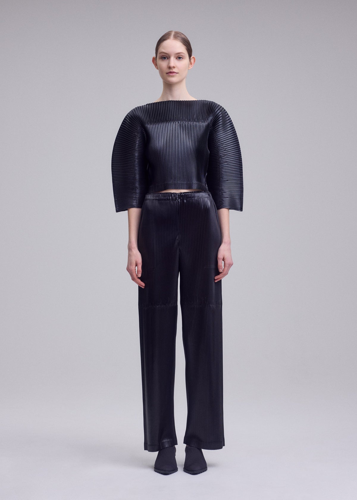 A model wears the ISSEY MIYAKE GLOSSED PANELS PLEATS glossed-panels-pleats-cropped-top-grey.
