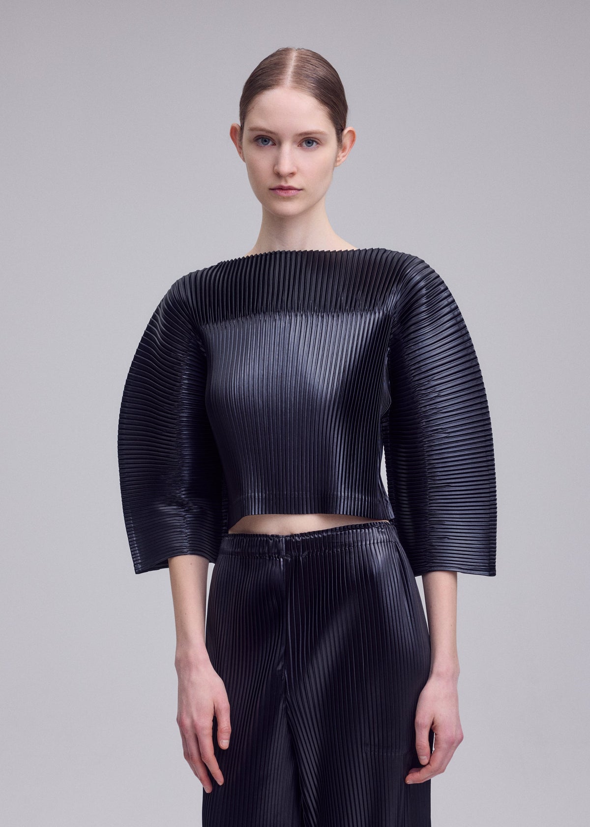 A model wears the ISSEY MIYAKE GLOSSED PANELS PLEATS glossed-panels-pleats-cropped-top-light-pink.