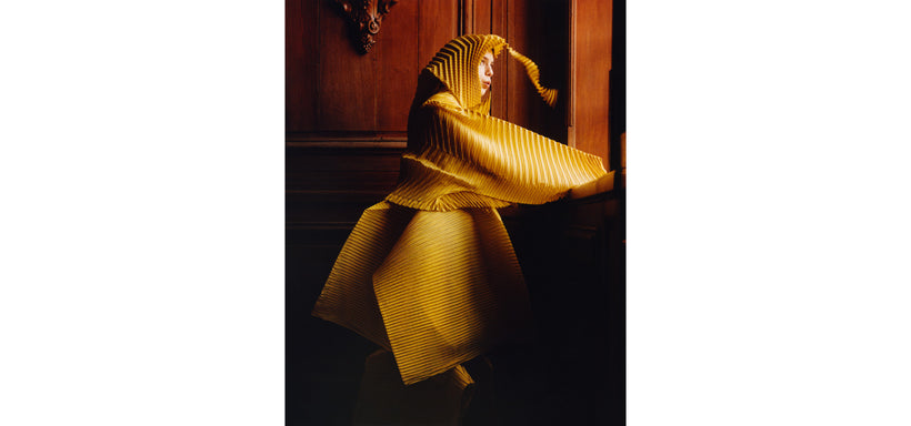 Centre: Model wearing GENERIC STRIPES (HAND PLEATS) Jacket in Yellow-Hued and GENERIC STRIPES (MACHINE PLEATS) Dress Yellow-Hued, set against a brown wooden background, looking out of the window.