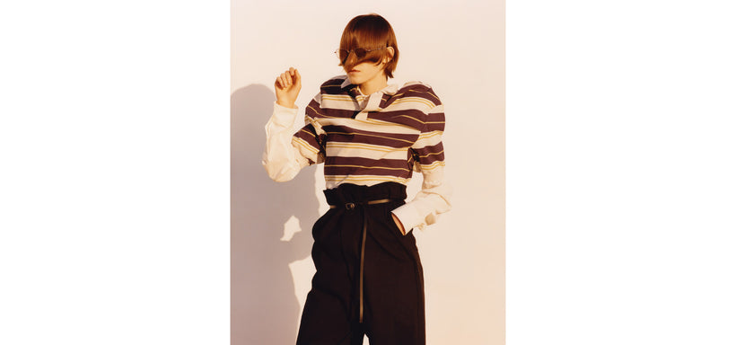 Centre: Model wearing GENERIC WEAR: STRIPES Top in Purple-Hued and GENERIC WEAR Trousers in Black, confident pose against a light pink background.