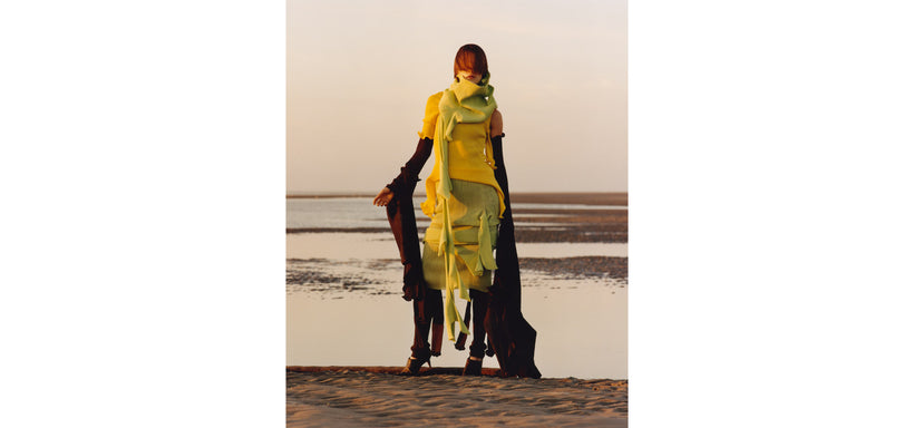 Centre: Model wearing ADVENTITIOUS: SLEEVES Top and Dress in Parakeet Yellow, in an expressive pose against a seascape background.