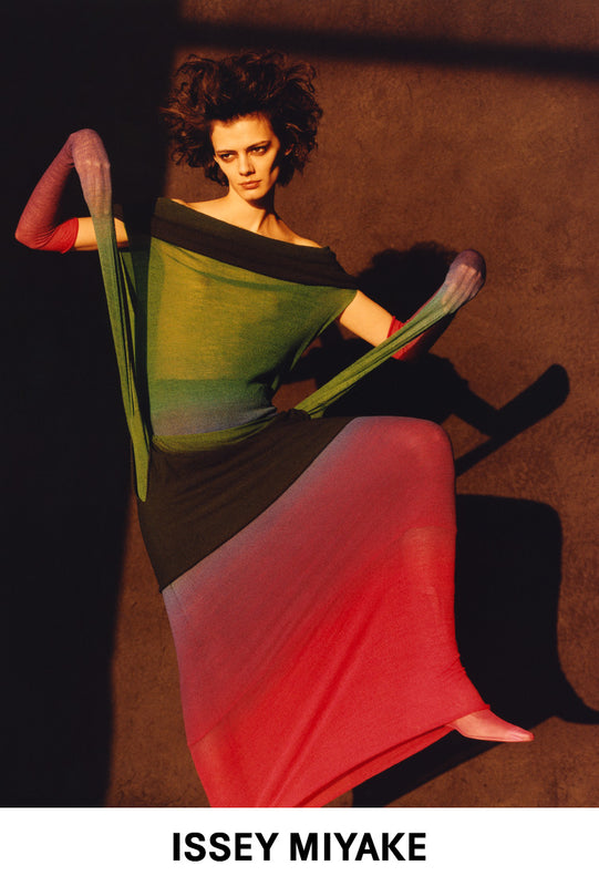 Main: Model wearing NUANCE TUBE Skirt in Afternoon as a dress, dynamic pose with left knee in the air. Yellow lighting. Brown background. Bottom centre: ISSEY MIYAKE logo in black on a white background.