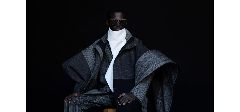 Centre: Model wearing KASURI Coat and Trousers in Black and IM-101 Glasses in Silver Matte, sitting on a wooden stool and looking straight ahead against a black background.