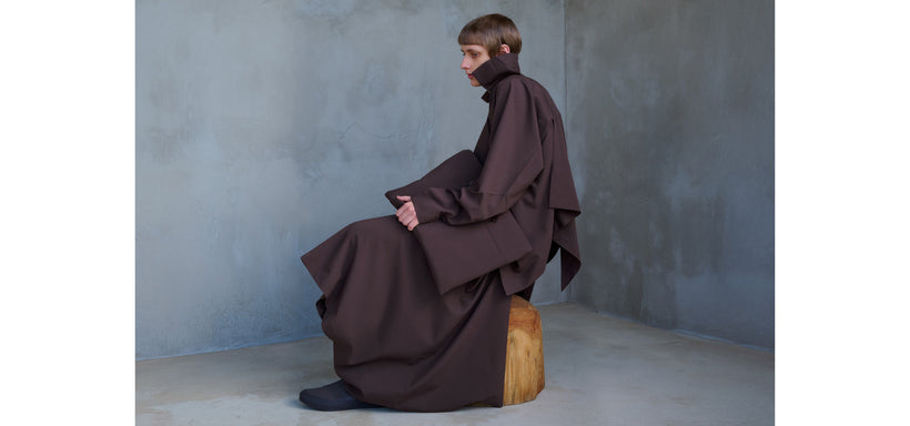 Centre: Model wearing FLAT DRAPE Jacket in Dark Purple, seated on a wooden stool against a grey concrete background.